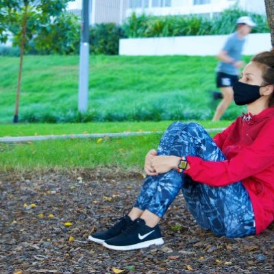 Woman sitting against tree with facemask on.